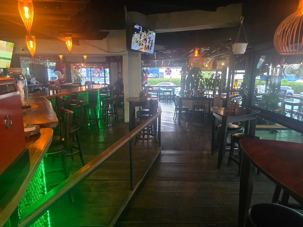 Barra de madera del bar con luz verde bajo el mostrador, bancos altos, un televisor encendido y mesas junto a los ventanales que dan a la calle.
