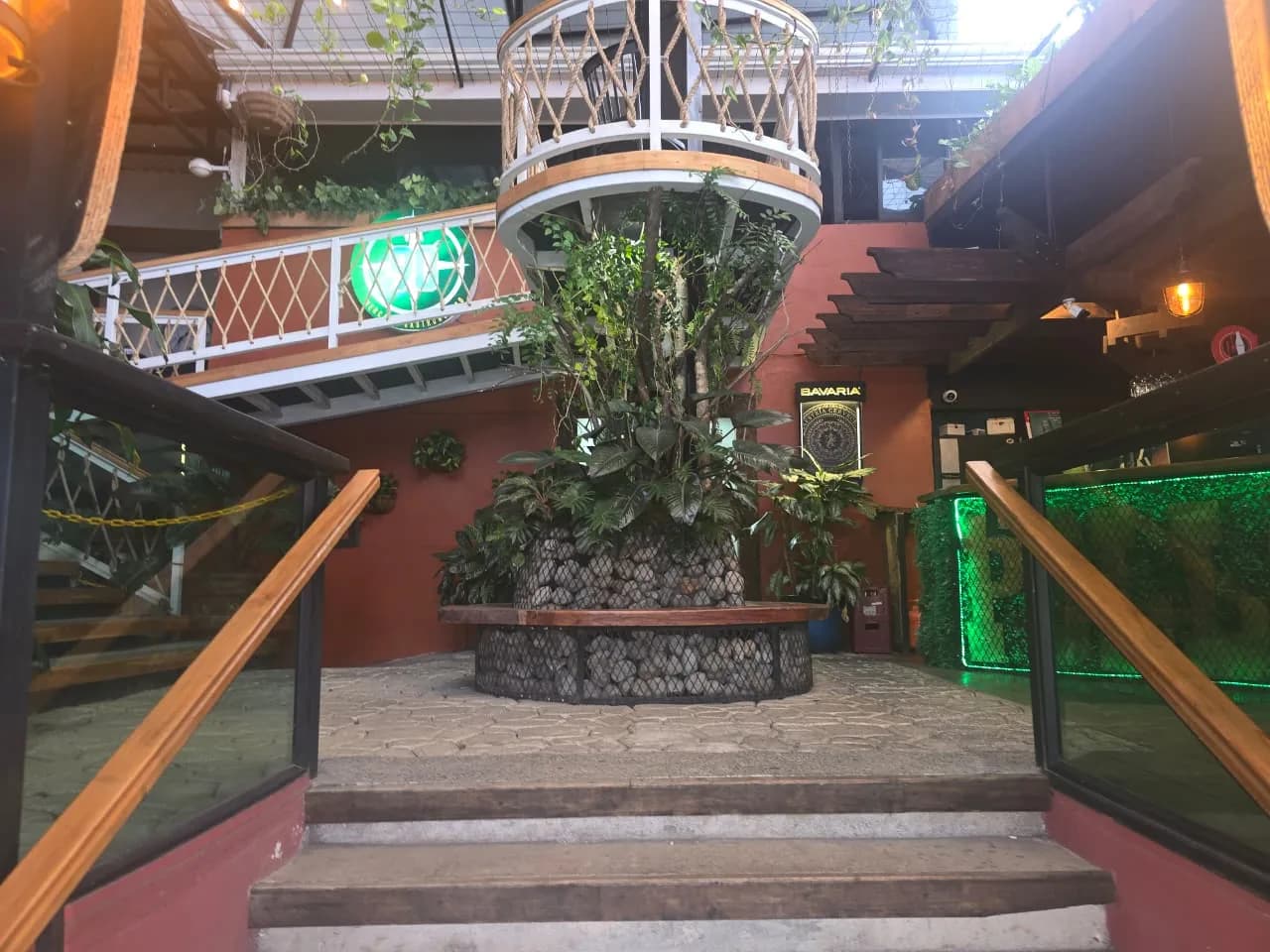 Patio central del mercado: una jardinera circular de piedra con banca de madera alrededor de un árbol, el balcón redondo del segundo piso arriba y el rótulo verde de Vía 90 al costado.