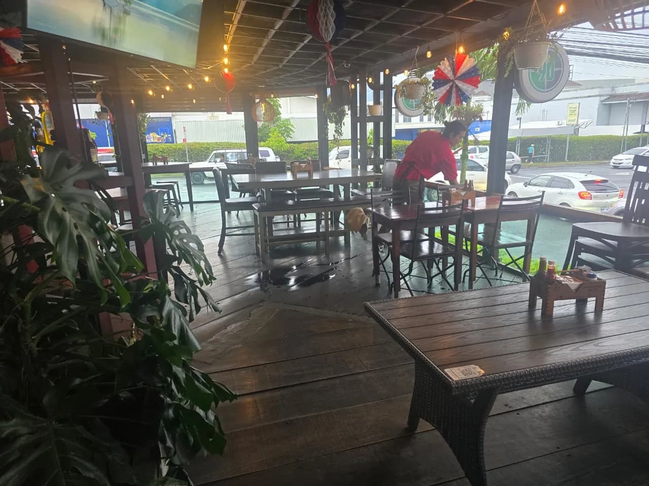 Terraza techada con mesas de madera y decoración de banderines azul, blanco y rojo; una persona con camisa roja alista una mesa y al fondo se ve la calle.
