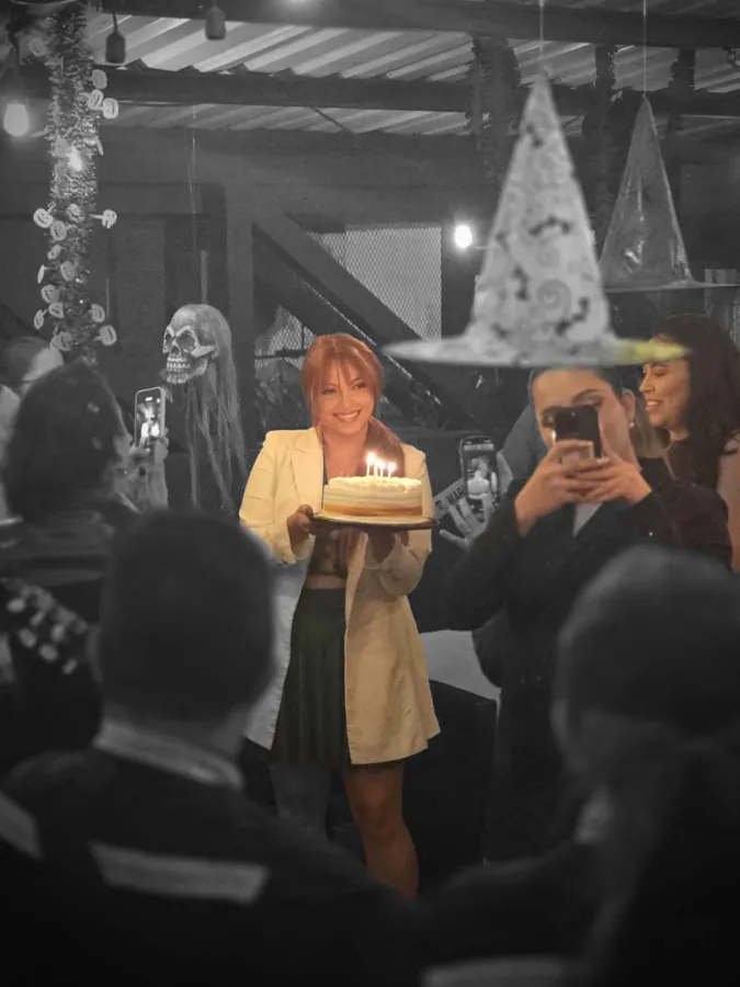 Una mujer camina sonriendo con un queque de cumpleaños encendido entre la gente, que la graba con los celulares, en medio de la decoración de Halloween del local.