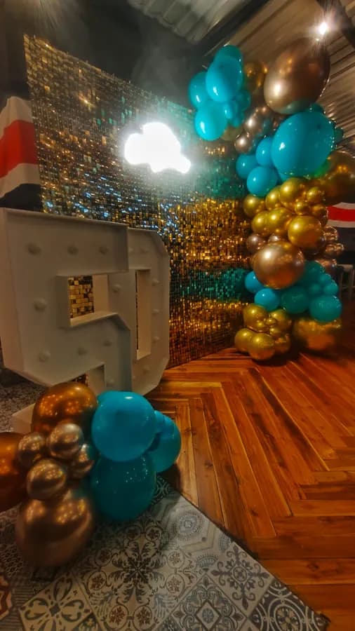 Rincón de fotos montado para una fiesta: una pared de lentejuelas doradas, un arco de globos turquesa y dorados y una letra gigante iluminada sobre el piso de madera.