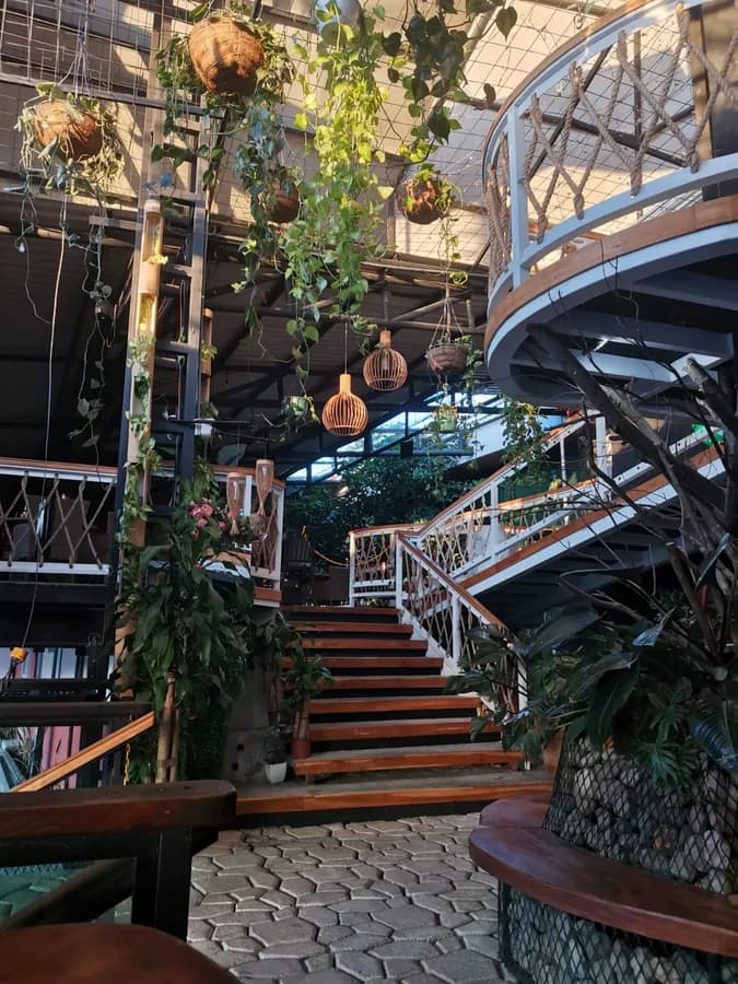 Atrio del mercado: la escalera de madera que sube al segundo piso, con lámparas de mimbre colgantes, plantas cayendo desde la estructura metálica del techo y el balcón redondo con baranda de cuerda a la derecha.