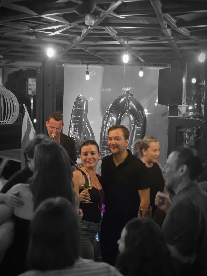 Gente conversando con copas en la mano en una fiesta de cumpleaños, con globos plateados en forma de número al fondo y un cantante con micrófono.