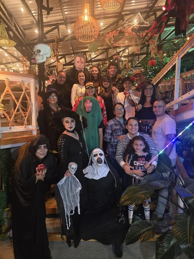Foto grupal de una fiesta de disfraces: alrededor de quince personas posan en las gradas de madera bajo las lámparas de mimbre, con telarañas y decoración de Halloween colgando del techo.