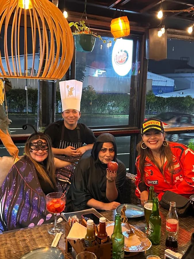 Cuatro personas disfrazadas comparten una mesa junto al ventanal, con bebidas y una lámpara de mimbre encendida encima; afuera se ve la calle de noche.