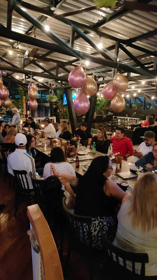 Una mesa larga llena de gente cenando bajo el techo de lámina y las luces colgantes, con racimos de globos rosados y dorados encima.