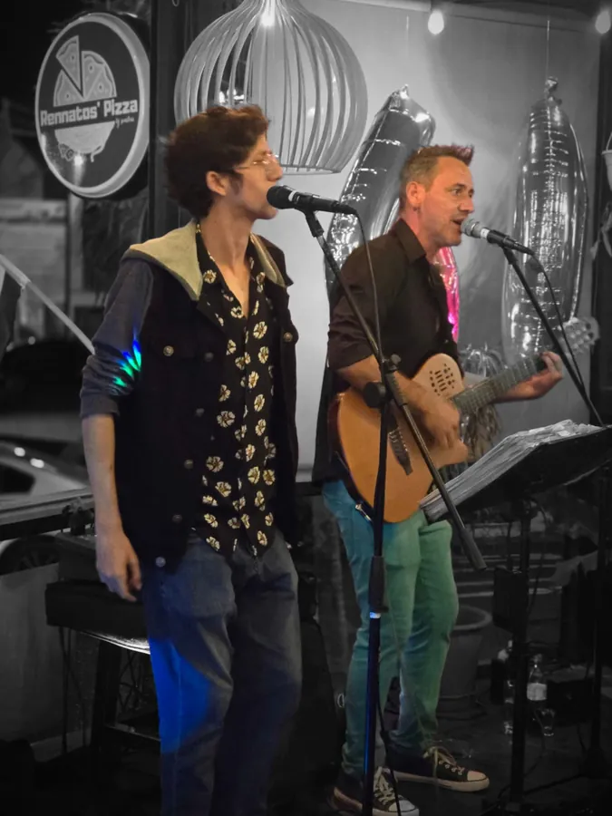 Dos músicos cantan frente a un micrófono, uno de ellos con guitarra acústica, junto al rótulo de Rennato's Pizza y globos plateados.