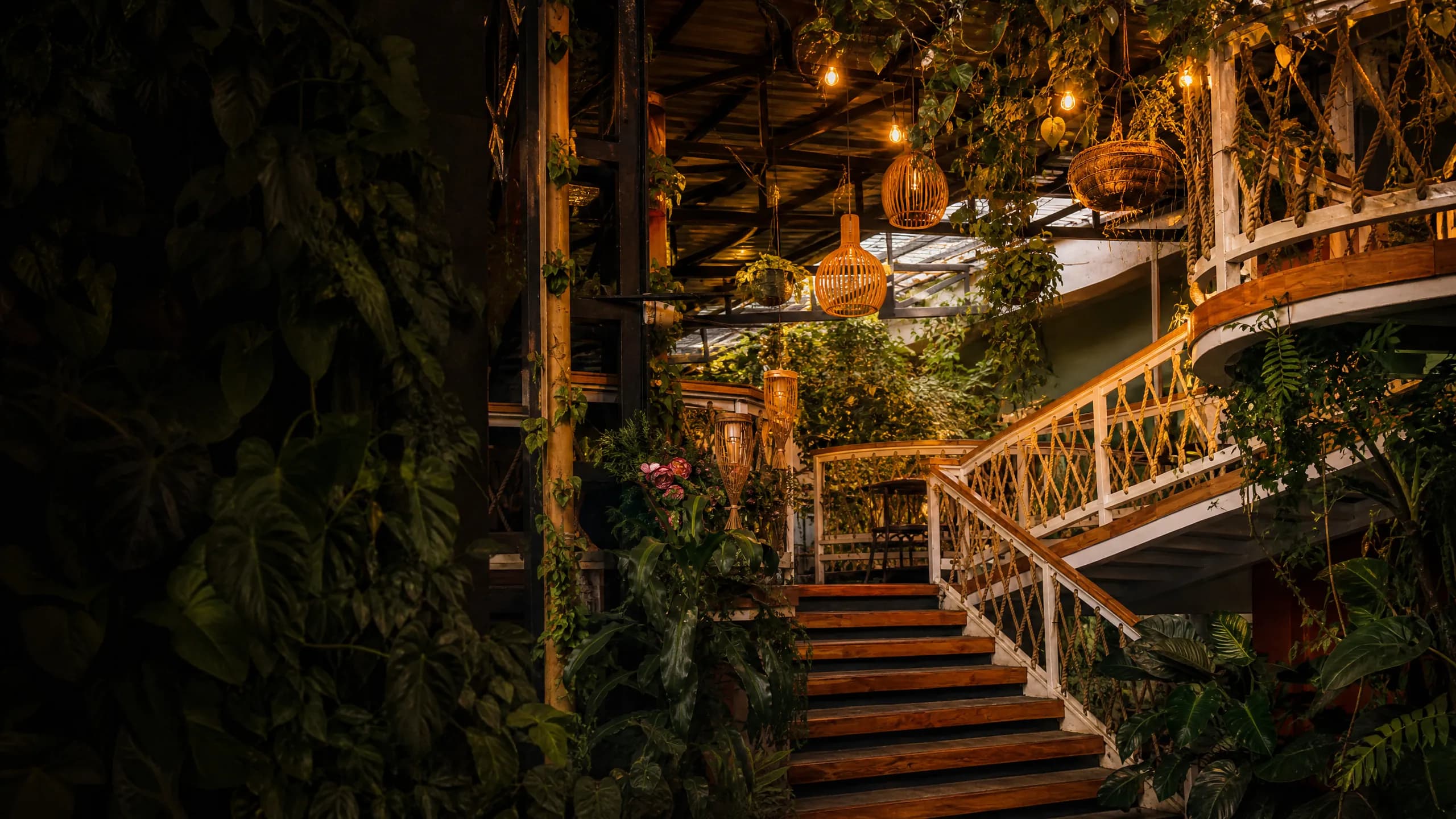 Atrio del mercado de noche: una escalera de madera sube entre barandas blancas tejidas con cuerda, con lámparas de mimbre encendidas, guirnaldas de bombillos y plantas colgando de la estructura del techo.
