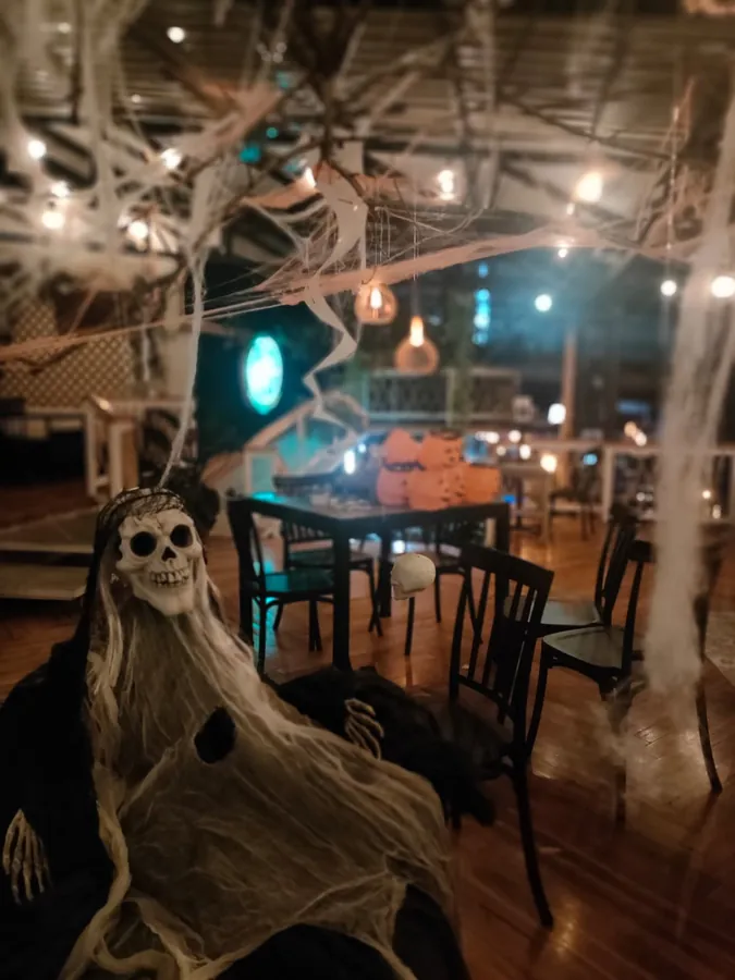 El salón decorado para Halloween y todavía vacío: telarañas colgando del techo, calabazas sobre una mesa, un esqueleto en primer plano y bombillos encendidos sobre el piso de madera.
